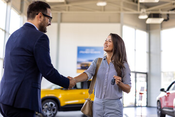 Naklejka premium Smiling salesman or manager giving car keys and shakes hand to modern happy woman new car owner in car dealership