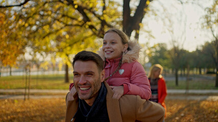 Fototapeta premium Father giving daughter a piggyback ride in a park on a sunny autumn day