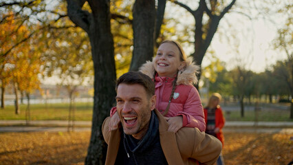 Fototapeta premium Father giving daughter a piggyback ride in a park on a sunny autumn day