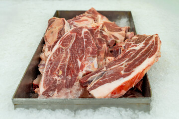 Thick, raw cuts of bone-in red meat, are displayed in a metal tray resting on a bed of crushed ice at a butcher shop or supermarket counter.