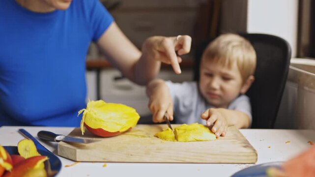 Mother teaching her young son to cut a mango fruit in the kitchen