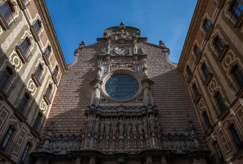 Facade of Montserrat Abbey, a Benedictine monastery in Catalonia, Spain. The Gothic and Renaissance architecture features a rose window, intricate carvings, and statues, creating a majestic scene