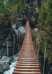 Fototapeta premium Wooden suspension bridge stretches over a rocky gorge surrounded by lush green trees in the mountains