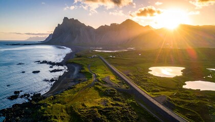 A dramatic coastal landscape with a winding road leading to jagged mountains, bordered by a black sand beach and lush green fields under a vibrant sunset sky.