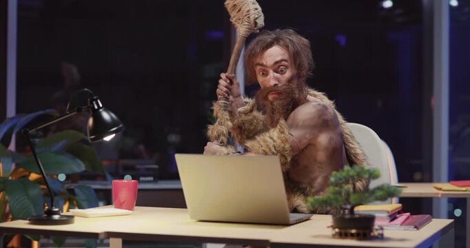 Prehistoric tribal savage cave human sitting by laptop computer interacting technology having fun screaming getting scared inside night office. Movie concept.
