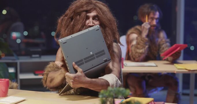Prehistoric tribal savage cave human sitting by laptop computer interacting technology having fun screaming getting scared inside night office. Movie concept.