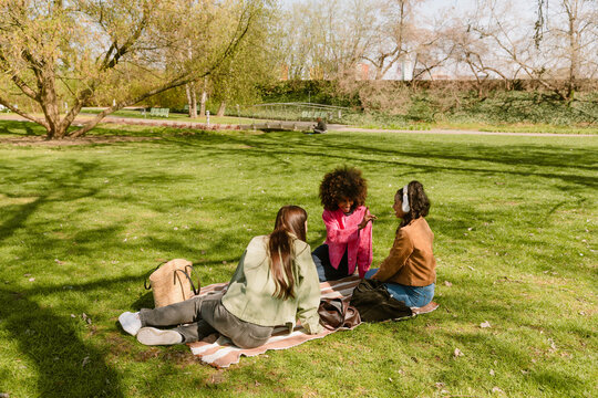 A woman shows her female friend who is listening to headphones while they laugh and sit on a blanket next to the female friend
