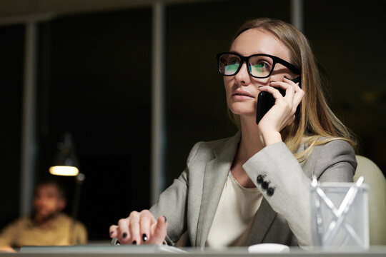 Attractive Caucasian businesswoman in elegant suit and glasses sitting at office table and talking on smartphone in the late evening