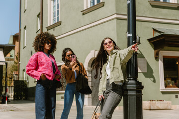 Obraz premium A woman leans on a street lamp and points to the side while laughing and standing next to her are two female friends, one of whom is holding a phone