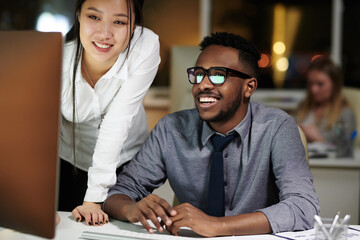 Cheerful colleagues working late in office. Young Asian woman and African American man looking at computer screen and smiling happily