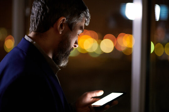 Caucasian middle-aged bearded businessman standing by office window in the late evening and using smartphone on blurred lights background - Powered by Adobe