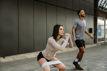 A female athlete squats with a rubber band on her thighs while a male athlete jumps on a rope next...