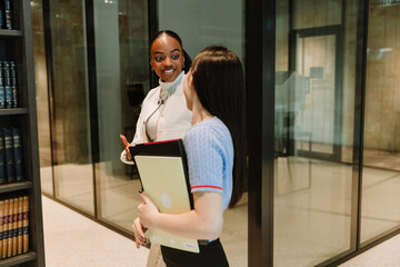 A female student is smiling and talking to a female classmate who is walking next to her and carrying a notebook and folder