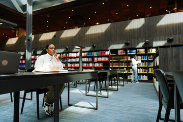 A female student looks thoughtfully in front of her and holds a pen over a notebook while sitting at a desk