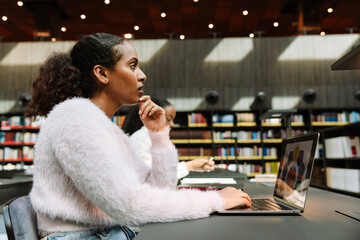 A female student looks thoughtfully in front of her while keeping her hand on a laptop and sitting...