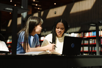 Two female students talking and sitting at a table while one of them holds a pen and the other looks at a laptop