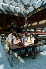 A female student stands and looks at a laptop with two other female students sitting at a table next to a female student reading a book