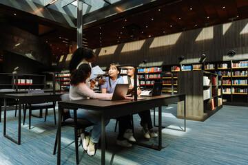 A group of three female students are sitting at a table and listening to a female classmate standing next to them holding a notebook and pen