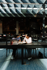 Two female students are talking and sitting at a table while one of them is holding a book and the other has her hands on a laptop
