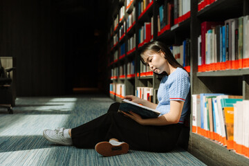 A female student is reading a book and sitting on the floor while leaning on bookshelves