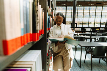 A female student is standing and leaning on bookshelves while reading a book