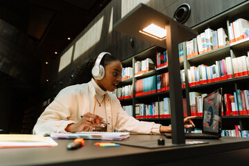 Fototapeta premium A female student pointing at a laptop and holding a pen while listening in headphones and sitting at desk