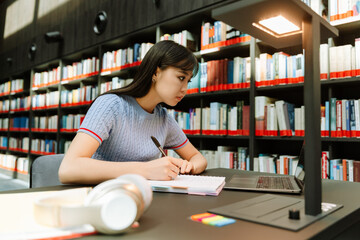 A female student is holding a pen over a notebook and looking at a laptop while sitting at a desk