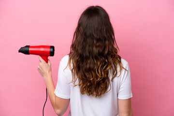 Young caucasian woman holding a hairdryer isolated on pink background in back position