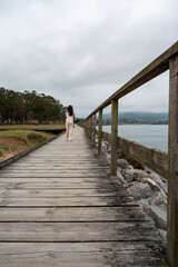 Fototapeta premium A woman is walking on a wooden bridge over a body of water. The sky is cloudy and the water is calm. The woman is alone and is looking out over the water