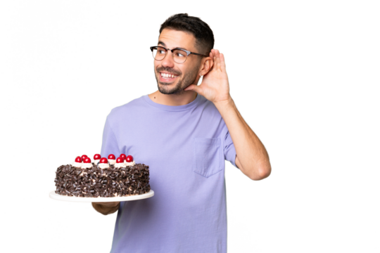 Young caucasian man holding birthday cake isolated on green chrome background listening to something by putting hand on the ear