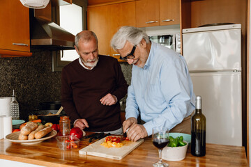 A man cuts tomatoes and smiles while standing at a table next to his friend and they talk