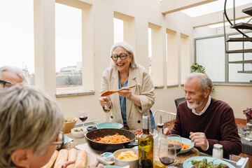 A woman holds a spatula and laughs while standing at a table with a group of three friends sitting around it