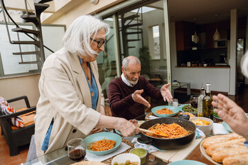 A woman stands and holds a spatula in spaghetti in a pan while two friends sit next to her