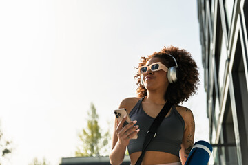Female athlete holding mat and phone while listening in headphones and looking away