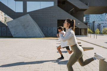 A woman and a man squat on one leg while leaning their other legs on stone chairs