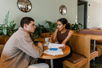 A female employee holds documents and talks to a male employee sitting across from her at a desk