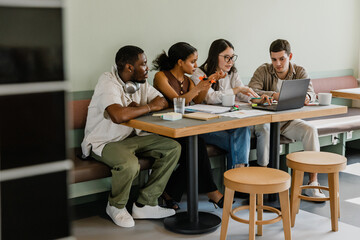 A group of four workers are sitting at a table and talking while one of them is typing on a laptop...