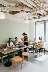 A female employee shows a notebook to two employees who are sitting at a table while a male employee is typing on a laptop next to them