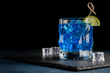 Colorful blue cocktail in a glass with a slice of lime on a black background