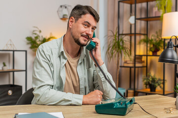 Caucasian man freelancer working, talking with client on retro old-fashioned wired telephone at home office table. Salesman in casual clothes. Remote distant job. Employment occupation. Lifestyle.