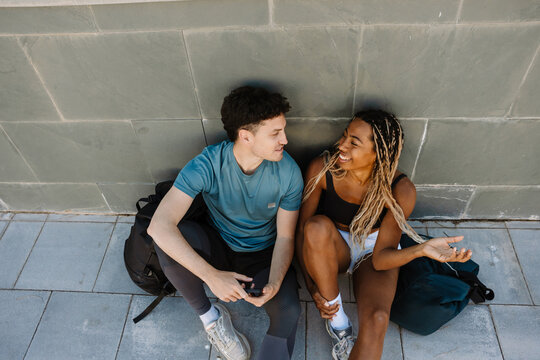 Female athlete laughing and listening to male athlete holding a phone while they sit on the ground and lean on a wall - Powered by Adobe