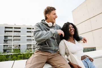 A female student stands and leans on the wall on which a male student is sitting, who hugs her while they laugh
