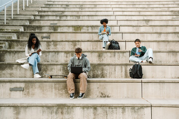 A group of four students sit on bleachers and read books and notebooks and look at a laptop
