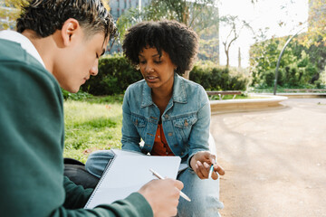 A female student is talking to a male student sitting next to her while they hold notebooks and pens