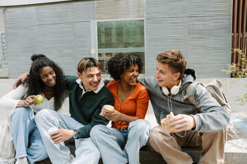 A group of four students sit on a curb and hug while laughing and eating