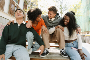 A group of four students laugh and lean on each other while sitting on a table and bench