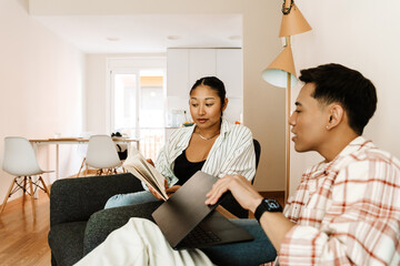 A man sits on a sofa and holds a laptop while reading a book shown by a woman sitting next to him on a chair
