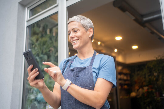 Mature florist using mobile phone to check flower orders outside her small business shop. - Powered by Adobe