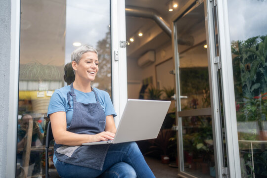 Mature flower shop owner sitting in front of the flower shop using laptop.