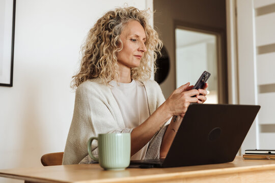 The mother is typing on the phone and sitting at the table in front of the laptop